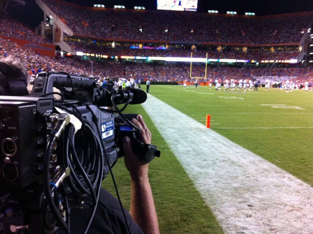 Camera operator filming live football game from the sidelines with stadium crowd in the background