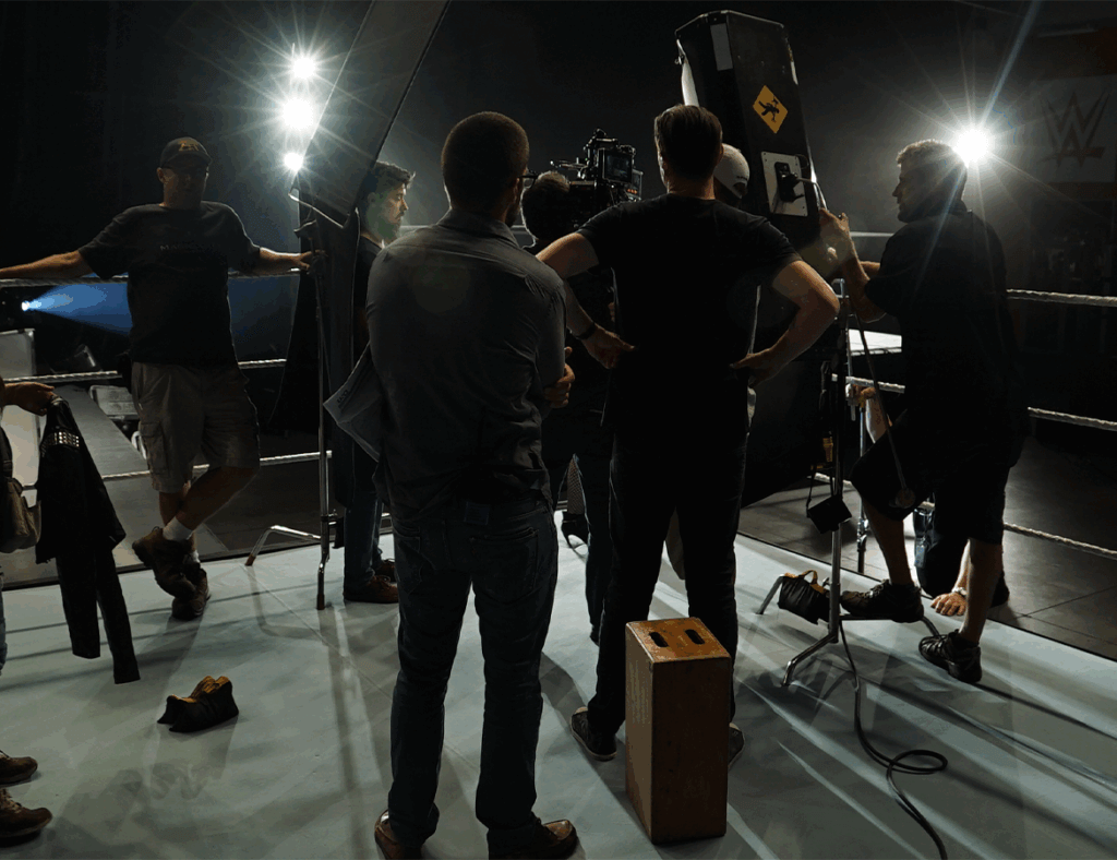 Full Size Team, Fractional Cost Image Showing: A film crew setting up equipment and observing an ongoing content shoot inside of a boxing ring.