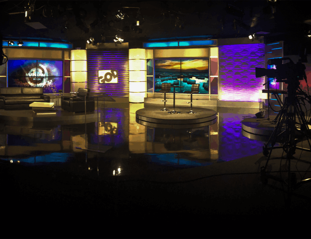 Image Showing: A vacant, dimly lit tv show set with purple and yellow branding. A video camera sits on a stand in the right corner of the image.